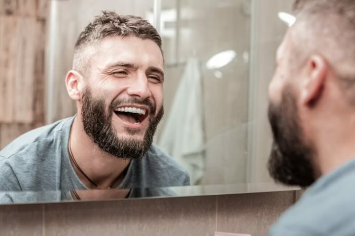 A smiling bearded man looking at himself in a bathroom mirror