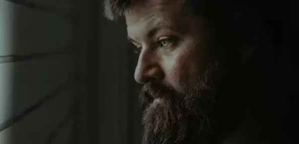 A bearded man looking out a dark window