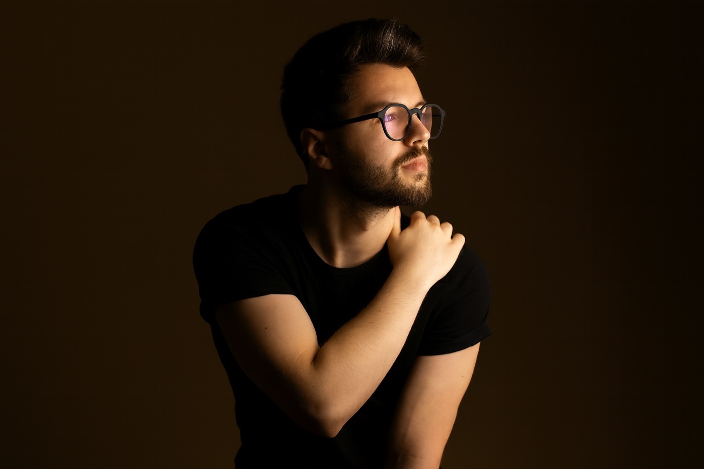 A man in glasses in low light, one hand on his shoulder, looking up
