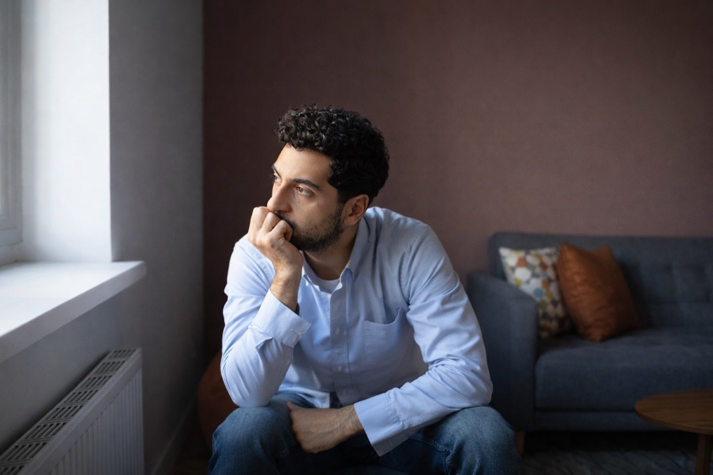 A man sitting by a window with his chin on his hand, deep in thought